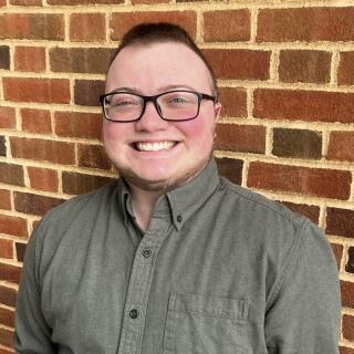 A masculine person with brown hair wearing a green shirt and black glasses against a brick wall