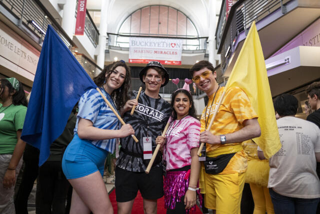 Four students in color team gear hold flags on the red carpet in front of the BuckeyeThon Banner