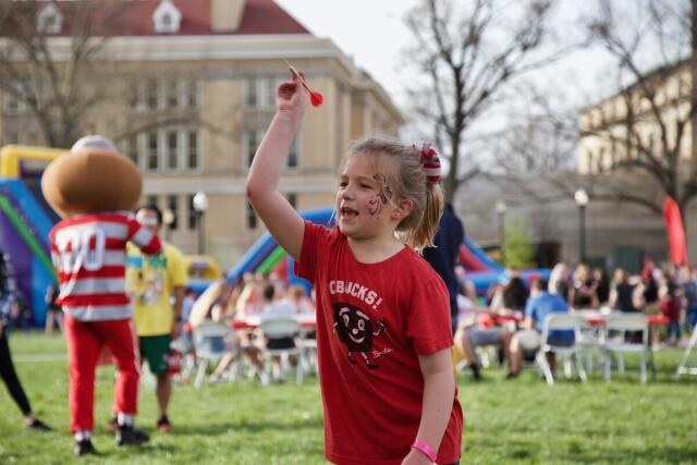 Girl in a red shirt reading "Go Bucks!" is about to through a dart at an event on the south oval.