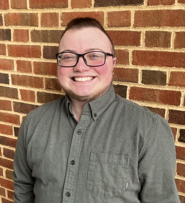 A masculine person with brown hair wearing a green shirt and black glasses against a brick wall