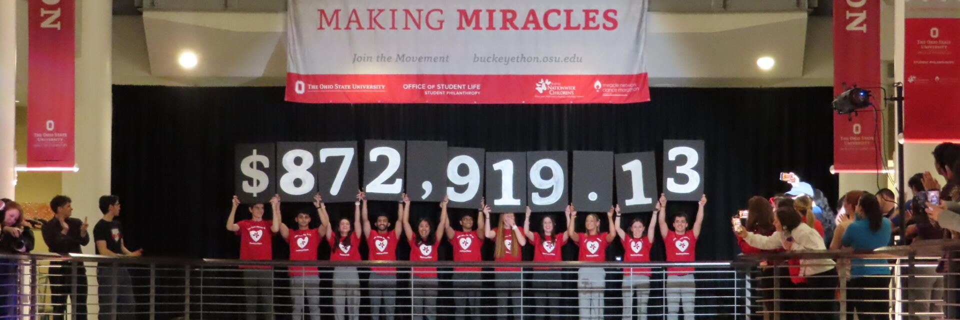 Several students facing a balcony where eleven students in black hold up signs showing $872,919.13.